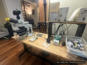 Home lab bench with an Olympus BX61 microscope, a Canon camera mounted on its camera port, a joystick and keypad, and the microscope control boxes on a shelf behind.